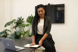 Naomi leans against a conference table in a meeting room, with a laptop in front of her and a tropical indoor plant behind her.
